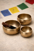 Load image into Gallery viewer, Three brass singing bowls on a textured surface with prayer flags in the background.