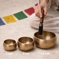 Load image into Gallery viewer, Three brass singing bowls with sizes labeled, being played on a mat with prayer flags.