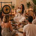 Load image into Gallery viewer, Person leading a meditation or sound bath session with participants in a room decorated with plants and musical instruments.