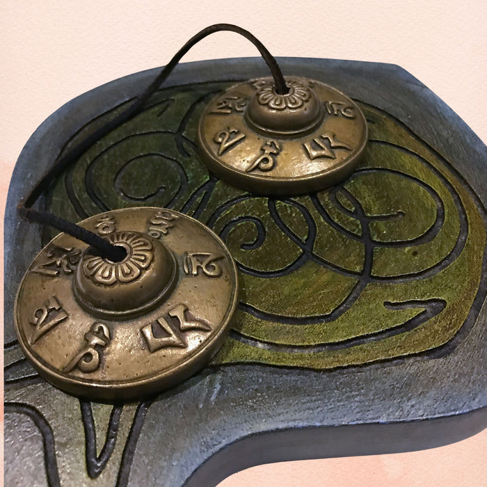 Mantra Tingsha cymbals with Tibetan symbols, aged brass finish and hanging cord on decorative cloth.